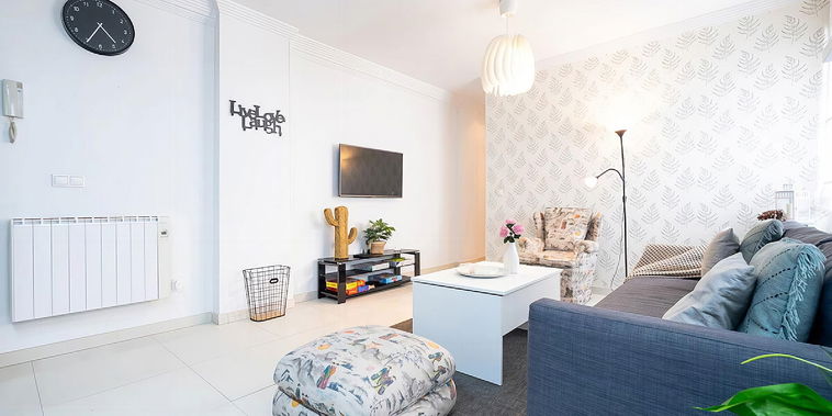 Communal lounge with TV, sofa, coffee table, armchair, ottoman, small chest of drawers with plants on top, wallpaper on the wall.