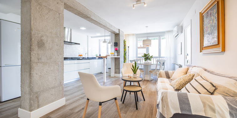 The living room opens to the kitchen with a concrete pillar, sofa, and large windows, a coffee table, and chairs. A dining table and chairs are located by the windows.
