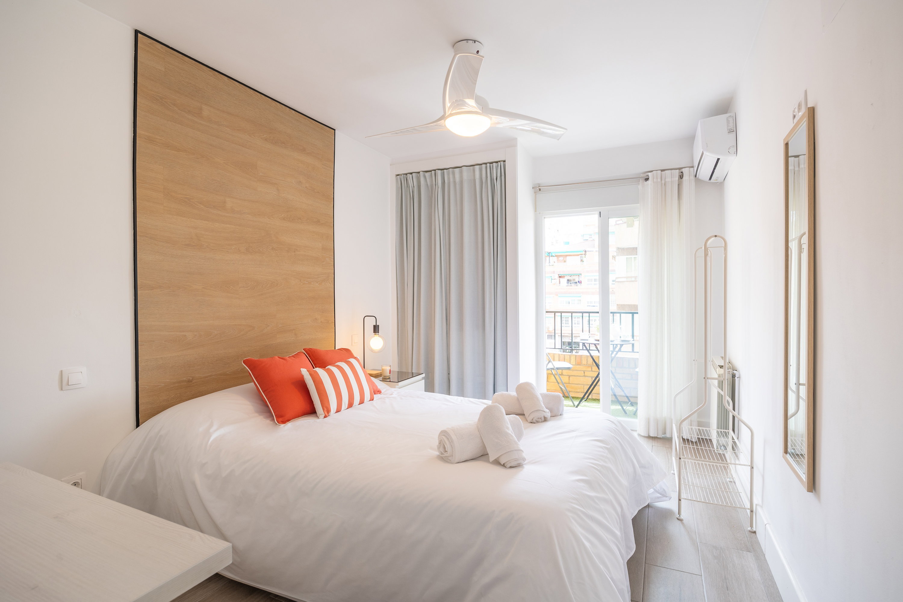 A bedroom, a fan, and access to the balcony. Mirrors line the walls,clothes rack, a shoe rack, set of towels, bedside table with a night lamp.